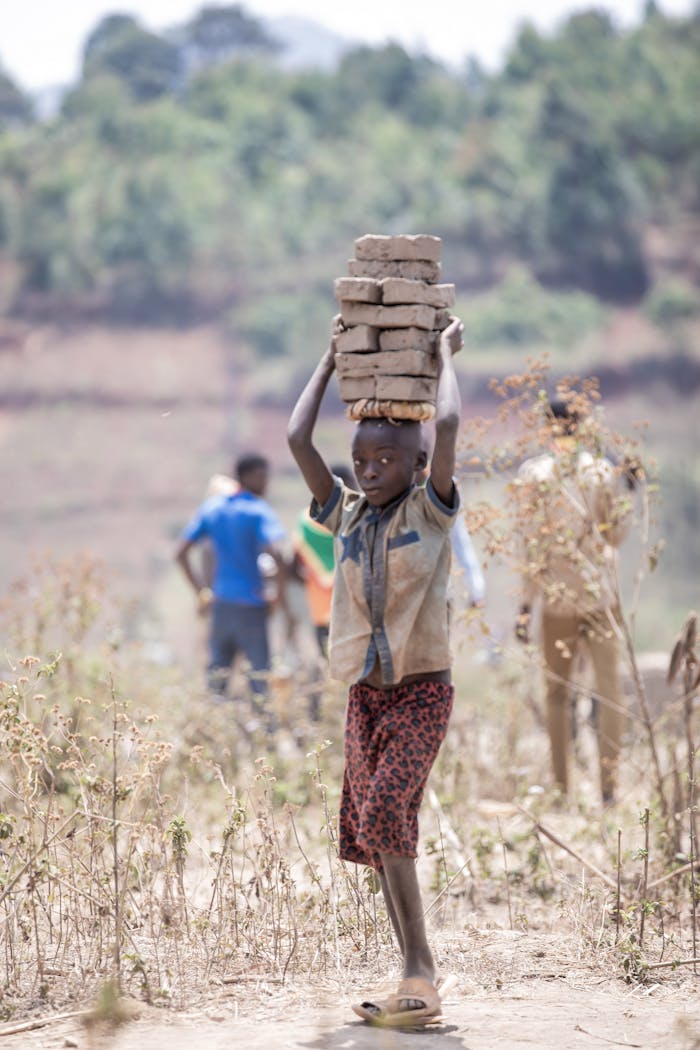 African child carries stack of bricks on head in rural area, underlining themes of labor and resilience.