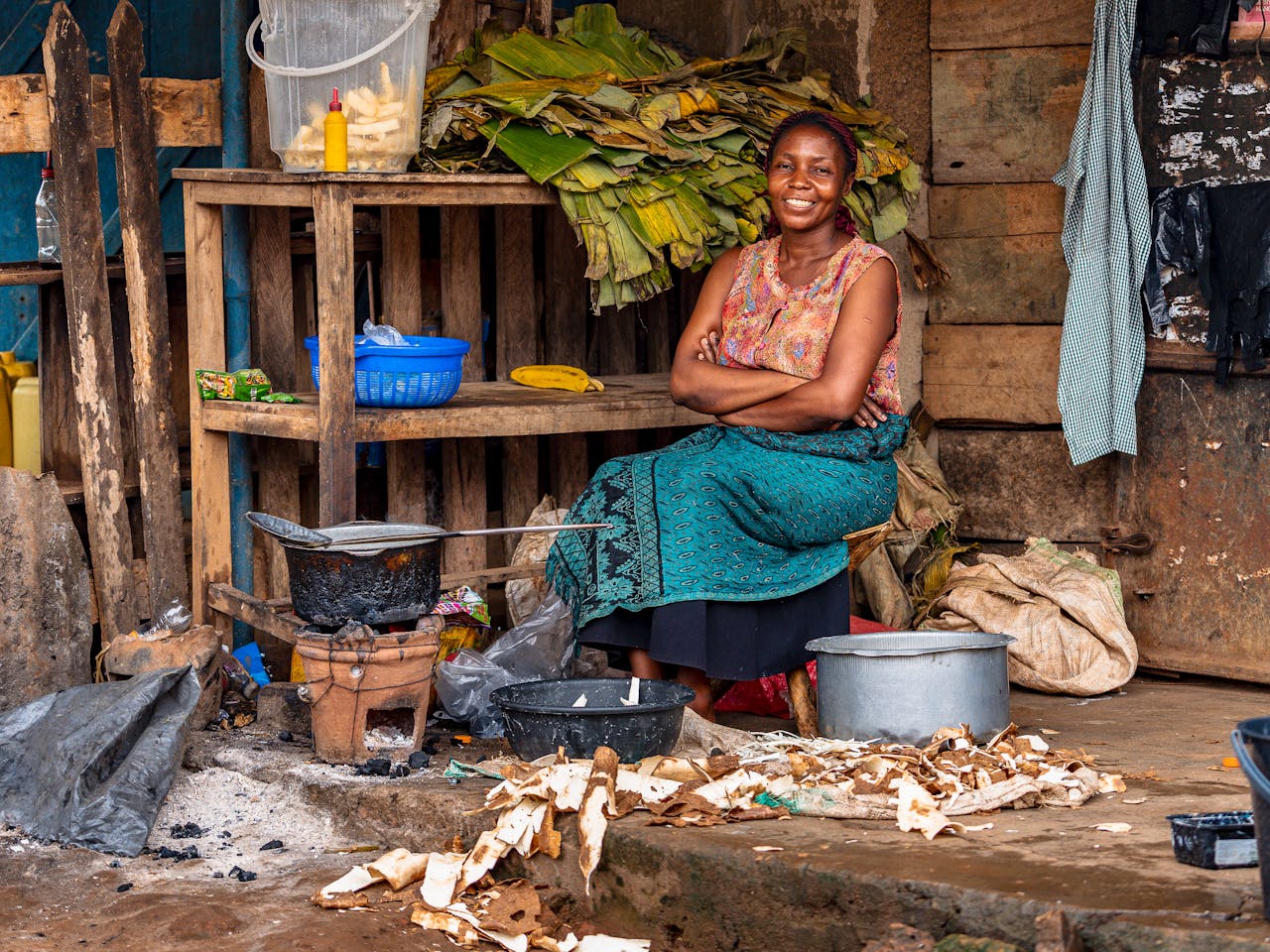 Smiling woman seated in an outdoor market stall surrounded by food preparation items.